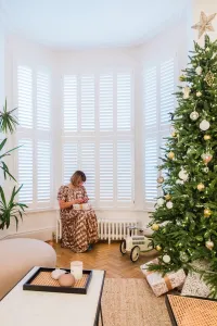 A low white Mercury 6 Column 360mm cast iron radiator under a shuttered bay window, with a woman wrapping gifts beside a Christmas tree.