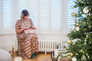 A low, plain white Mercury 6 Column 360mm cast iron radiator with a brass valve under a shuttered bay window, beside a Christmas tree on parquet.
