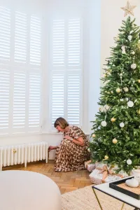 A low plain white Mercury cast iron radiator on feet under shuttered bay windows, a woman adjusting its valve beside a Christmas tree.