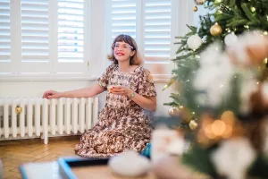 A low white Mercury 6 Column cast iron radiator on feet under shuttered windows, a woman in a sequin dress beside it and a Christmas tree.