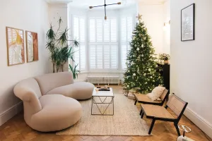 Low white Mercury cast iron radiator beneath a shuttered bay window, with a curved cream sofa, Christmas tree and herringbone floor.