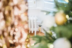 A low white plain Mercury cast iron radiator with a brass valve on a herringbone floor below a shuttered window, seen through Christmas tree branches.