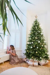 Low white Mercury 6 Column cast iron radiator on feet under a shuttered bay window, a woman seated by a decorated Christmas tree on parquet.