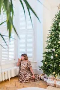 Low white Mercury cast iron radiator on feet with a brass valve under shuttered bay windows, beside a Christmas tree on a herringbone floor.