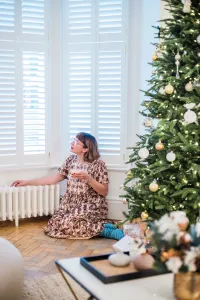 Low white Mercury cast iron radiator on feet under a shuttered bay window, a woman kneeling on parquet beside a decorated Christmas tree.