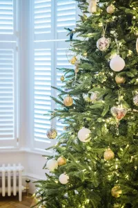 A decorated Christmas tree in front of white shutters, with a low white Mercury cast iron radiator and brass valve on a wooden floor behind.