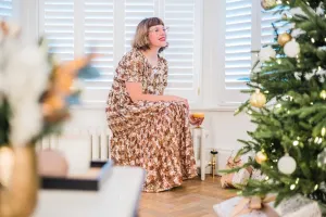 A woman in a sequinned dress sits by a white Mercury 6 Column 360mm radiator with a brass valve under shuttered windows, beside a Christmas tree.