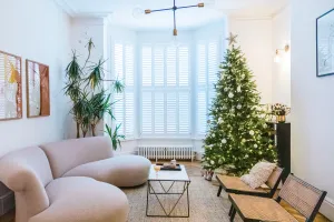 A low white Mercury 6 Column cast iron radiator under a shuttered bay window, beside a Christmas tree, curved sofa and cane chairs.