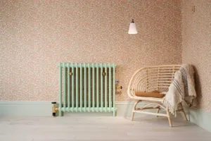 Plain Mercury 2 Column cast iron radiator in Little Greene Pea Green 91 on feet, brass valves, against pink floral wallpaper beside a rattan chair.