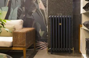 Plain Mercury 3 Column cast iron radiator in Midnight Ink on feet, with brass valves, against a speckled wall beside a wicker sofa.
