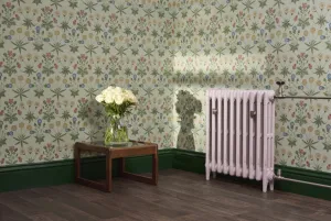 A plain Mercury cast iron radiator on feet in pale pink with Satin Nickel valves, against floral wallpaper beside a side table with white roses.