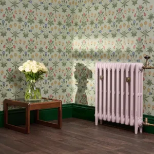 Pale pink Mercury 3 column cast iron radiator on feet with brass valves and wall stays, against floral wallpaper and green skirting.