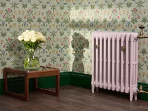Pale pink Mercury column radiator on feet with Natural Brass valves and wall stays, beside floral wallpaper and a table of white roses.