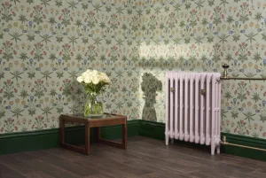 Plain pale pink Mercury 3 column radiator on feet with Natural Brass valves, beside a floral papered wall, dark green skirting and a table of white roses.
