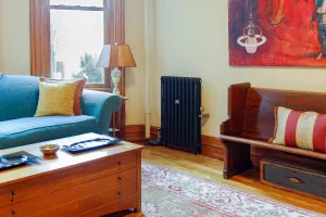 Plain Mercury 3 Column cast iron radiator in Matt Black on feet with valves, between a teal sofa and a wooden pew in a living room.