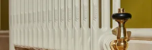 Close-up of a plain Mercury cast iron radiator in Farrow & Ball Lime White No. 1 with a polished brass Windsor angled thermostatic valve, against an olive wall.