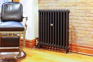 Mercury 3 Column 860mm cast iron radiator in Matt Black on feet against exposed brick, with brass valves, beside a vintage barber's chair.