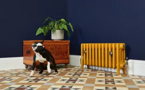 Low Mercury 4 column cast iron radiator in mustard yellow on feet with a nickel valve, against a navy wall on a geometric tiled floor with a dog.