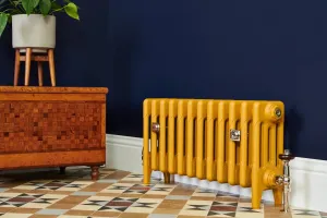 Low Mercury 4 Column 360mm cast iron radiator in mustard yellow on feet, with a nickel Windsor valve, against a navy wall on patterned tiles.