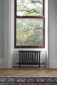 A low plain Mercury cast iron radiator in Astral Grey Mid on feet beneath a sash window, with brass valves, above a herringbone floor and rug.