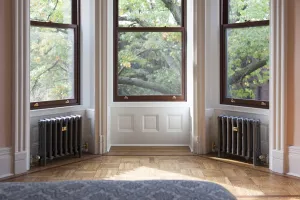 Two plain Mercury 4 Column cast iron radiators in Astral Grey Mid with brass valves, either side of a bay window above a parquet floor.