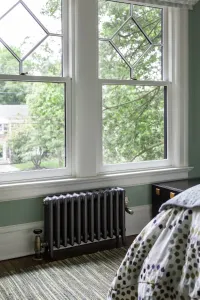 A plain Mercury 4 column cast iron radiator in Astral Grey Mid on feet beneath a sash window, with brass valves, in a green-walled bedroom.