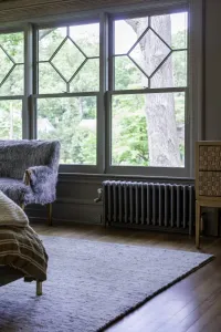 Low Mercury 4 Column cast iron radiator in Astral Grey Mid with a brass valve, wall-hung beneath leaded windows in a bedroom with a grey rug.