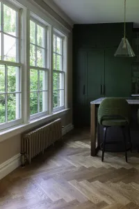 A low plain Mercury 4 Column cast iron radiator in Bronze Gold on feet beneath tall sash windows, by a herringbone oak floor and green kitchen.