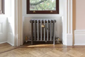 Plain Mercury 4 Column cast iron radiator in Astral Grey Mid on feet, with brass valves, in a white-panelled window recess above parquet flooring.