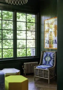 A low Mercury 4 Column cast iron radiator in Bronze Gold beneath a window in a dark green panelled room, beside a rattan chair and yellow pouffes.