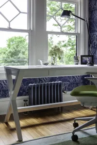A Mercury 4 Column 460mm cast iron radiator in Caspian on feet beneath a white desk by a bay window, with navy floral wallpaper and wooden floor.