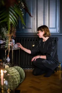 A woman kneels by a plain Mercury 4 Column cast iron radiator in Matt Black, beside a drinks trolley on a herringbone parquet floor.