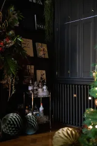 Plain Mercury 4 Column cast iron radiator in Matt Black beneath dark panelled cupboards, beside a brass drinks trolley and Christmas tree.