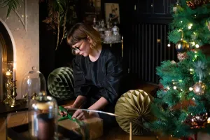 A woman wraps a gift by a Christmas tree, with a plain Matt Black Mercury cast iron radiator on the dark wall behind her.