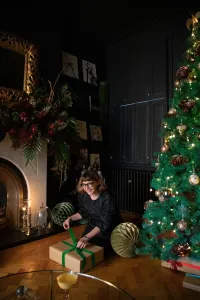 Plain Mercury cast iron radiator in Matt Black under a dark panelled window, beside a Christmas tree and a woman wrapping a gift on parquet.