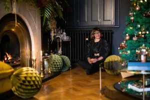 A plain black Mercury cast iron radiator below dark cabinets, with a woman seated on a parquet floor beside a fireplace and Christmas tree.