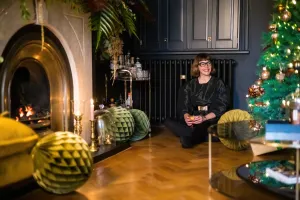 Plain Mercury cast iron radiator in Matt Black beneath dark cupboards, with a woman seated on a parquet floor beside a Christmas tree and fireplace.