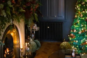 Plain Mercury 4 Column cast iron radiator in Matt Black on a dark wall beside a lit fireplace, drinks trolley and Christmas tree on parquet.