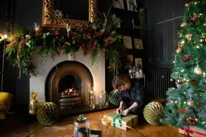 A plain Mercury cast iron radiator in Matt Black against a dark panelled wall beside a Christmas tree, a marble fireplace and a woman wrapping a gift.