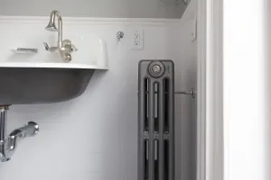 Plain four-column Mercury cast iron radiator in Astral Grey Mid with a wall stay, beside a wall-hung basin on white tiles.