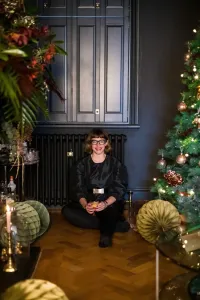 A plain Mercury cast iron radiator in Matt Black under a shuttered window, a woman seated on herringbone parquet beside a Christmas tree.