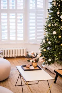 Low white Mercury 6 Column cast iron radiator on feet beneath a shuttered bay window, with a Christmas tree and marble coffee table on herringbone floor.