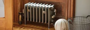 Low Mercury 6 Column cast iron radiator on feet in a metallic pewter finish with brass valves, in a wood-panelled alcove on parquet.