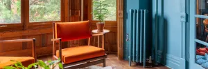 Plain Mercury cast iron radiator on feet in Farrow & Ball Stone Blue No. 86, with chrome valve, against a blue wall beside an orange armchair.