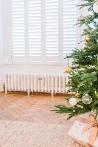 Low plain white Mercury 6 Column cast iron radiator on feet with brass valve, under shuttered window on herringbone floor beside a Christmas tree.