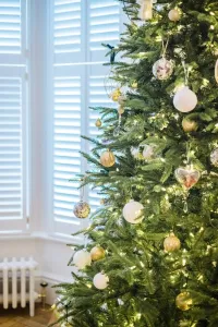 A white plain-column Mercury cast iron radiator with a brass valve sits below shuttered bay windows, partly hidden by a decorated Christmas tree.