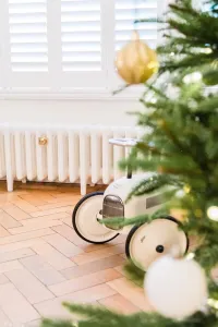 A low white Mercury 6 Column cast iron radiator on feet beneath shuttered windows, herringbone floor, toy car and Christmas tree in front.