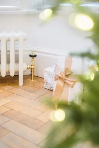 Plain off-white Mercury cast iron radiator on feet with a brass valve, on a herringbone wood floor beside a wrapped present and Christmas tree.