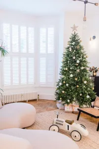 A low white Mercury 6 Column 360mm cast iron radiator on feet beneath a shuttered bay window, beside a decorated Christmas tree on parquet.