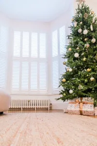 Low white Mercury 6 Column 360mm cast iron radiator on feet beneath shuttered bay windows, beside a decorated Christmas tree and wrapped gifts.
